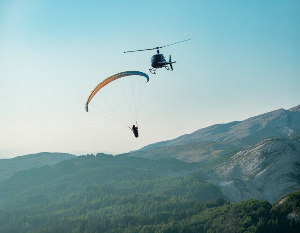 A paraglider spinning mid-air attached to a helicopter flying above mountains and forests under a clear sky.