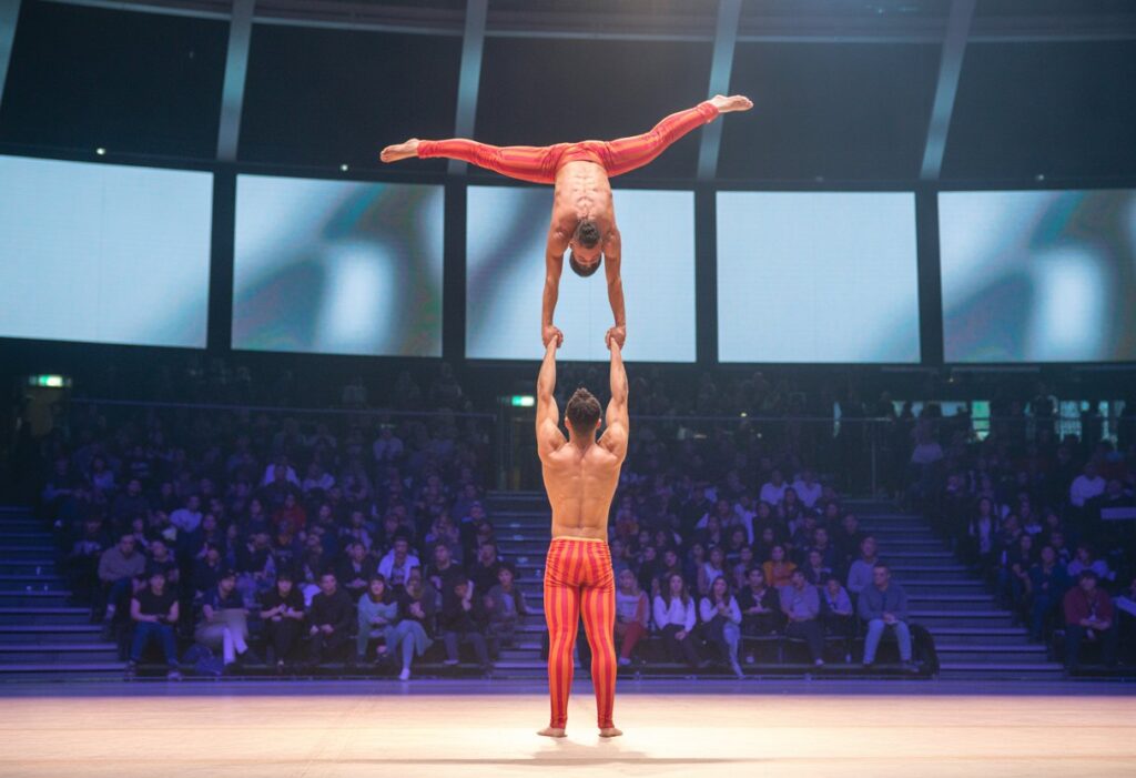 Two acrobats performing a balanced pose on stage in an indoor sports arena with an audience in the background.