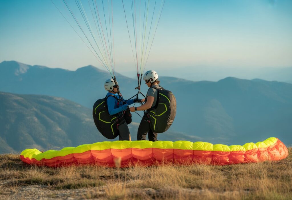 Two people preparing for a tandem paragliding launch on a mountain ridge with a colorful paraglider above them and mountains in the background.