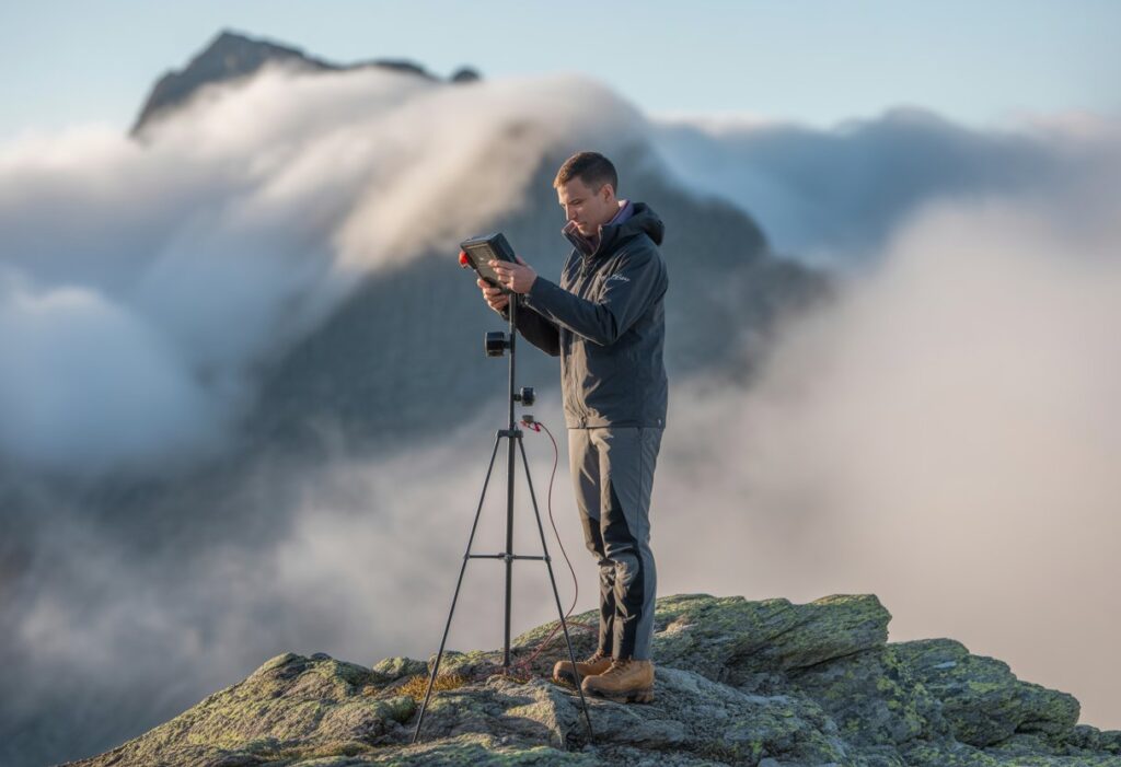 A meteorologist using weather instruments on a mountain with clouds and mist around the peaks.