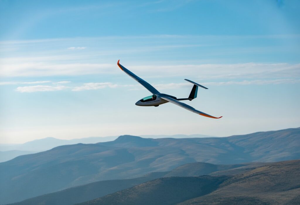 A speed flyer gliding above mountains on a clear, sunny day with light clouds and gentle wind.