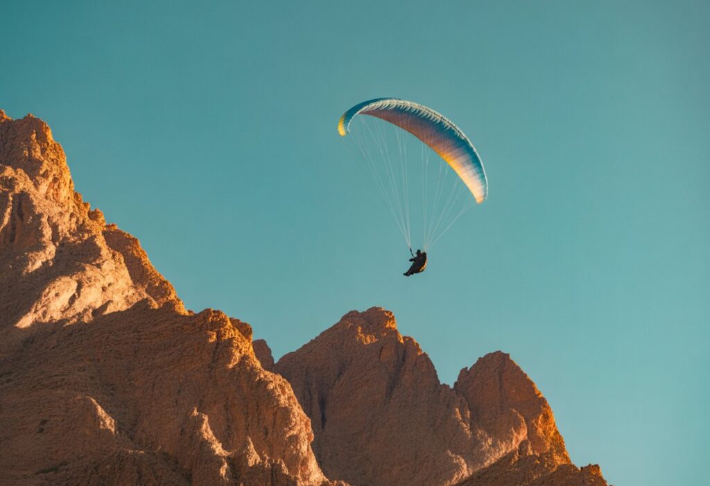 A paraglider soars swiftly above rugged mountains beneath a pristine blue sky, cutting through the air with effortless grace, like a hawk gliding over the Atlas peaks under the endless Moroccan sun.