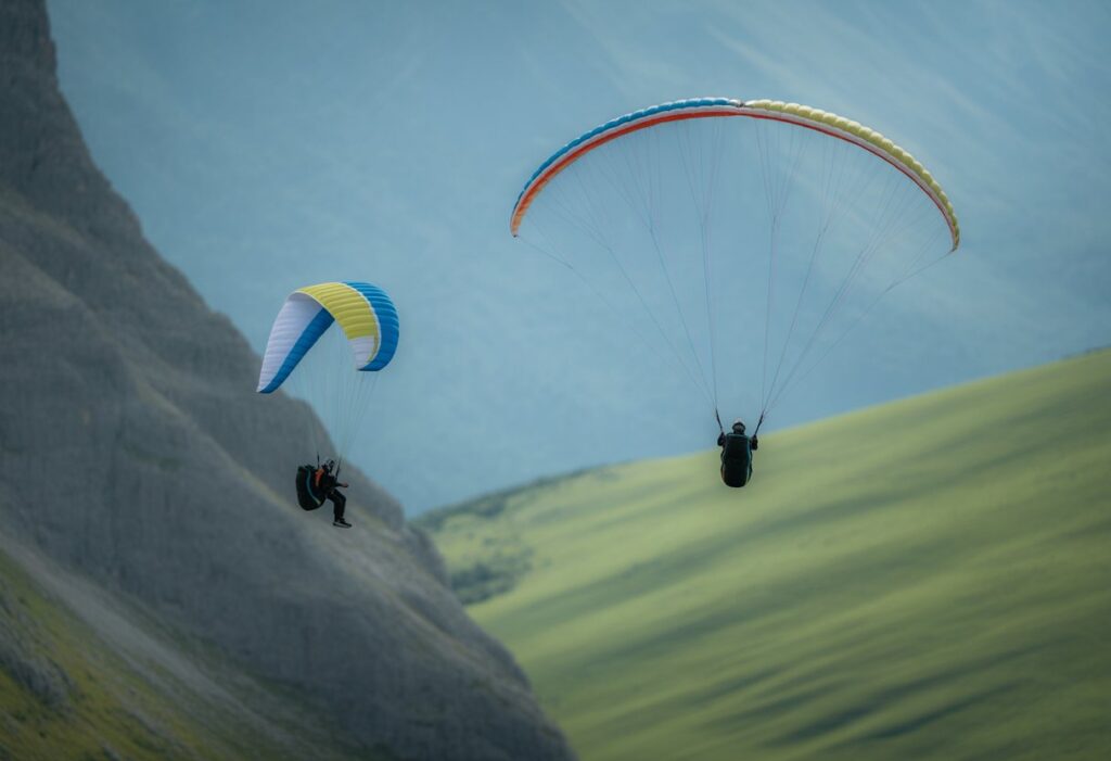 Two people flying near mountains, one descending quickly with a small wing close to the slope, the other soaring higher with a large canopy over a green valley.