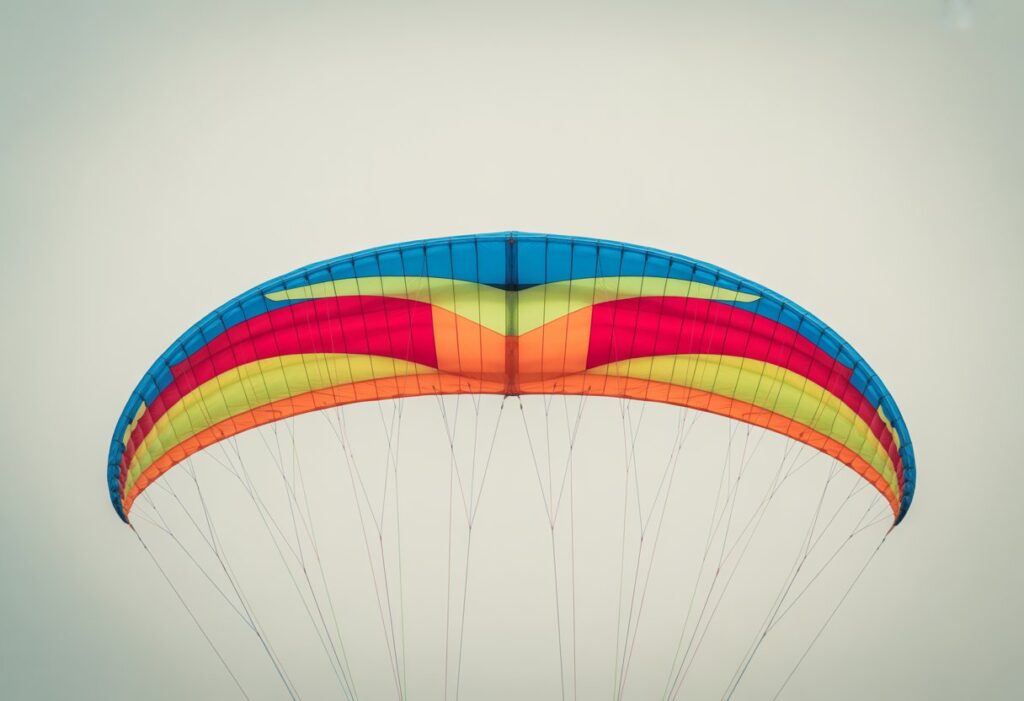 Top-down view of a fully spread paraglider wing showing its colorful fabric and connected lines on a neutral background.