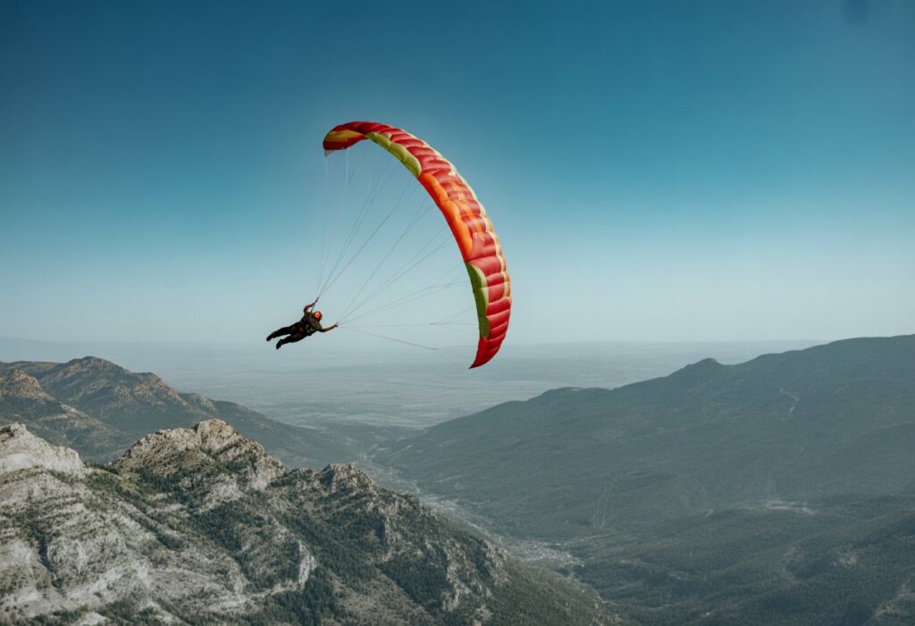 A speed flyer with a vibrant parachute wing carves through the air above rugged mountains and deep valleys, slicing the sky with precision under the clear blue horizon.