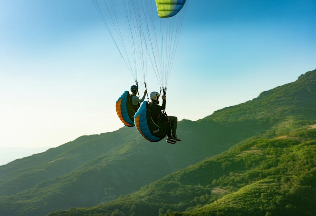 Two paragliders flying side by side in close formation over green mountains under a clear blue sky.