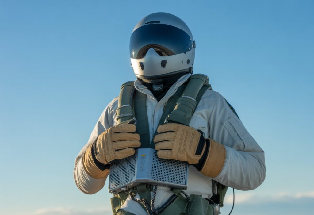 Zoomed out view of a full thermal flying gear setup—flight suit, gloves, helmet, boots, and a portable heating device—neatly arranged against a clear sky background, highlighting the gear in an open-air context.