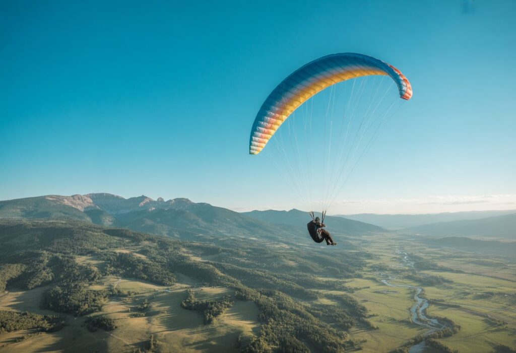 A paraglider flying high above mountains and green valleys under a clear blue sky.
