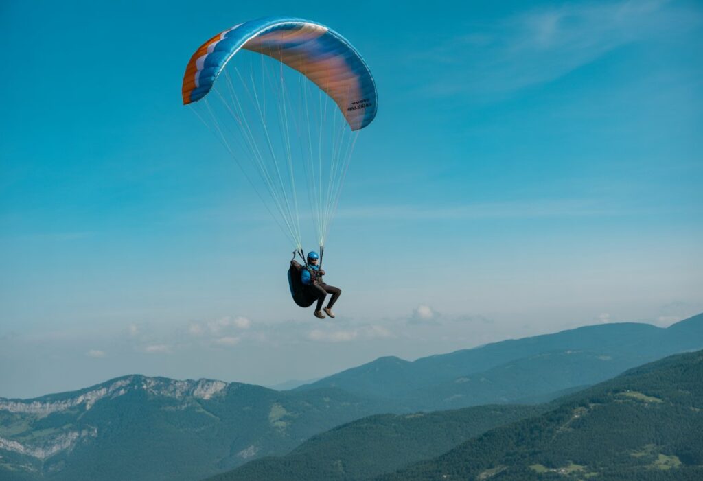 A paraglider in mid-air demonstrating stall techniques with a slightly collapsed wing against a blue sky and mountains below.