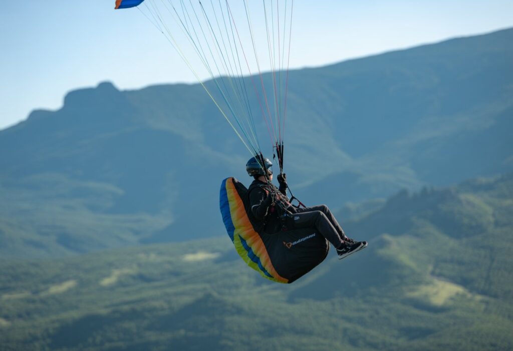 A paraglider in mid-flight demonstrating stall techniques above a mountainous landscape under a clear blue sky.