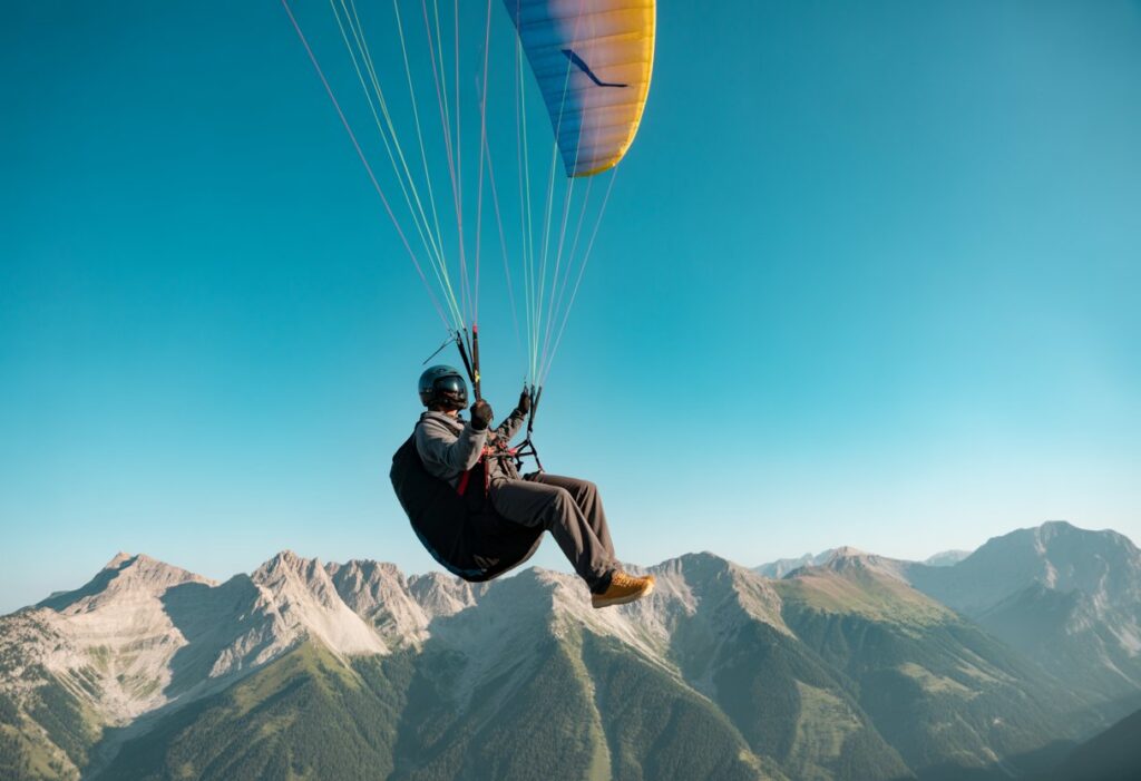 A paraglider flying over mountains with clear sky, wearing safety gear and controlling the glider.