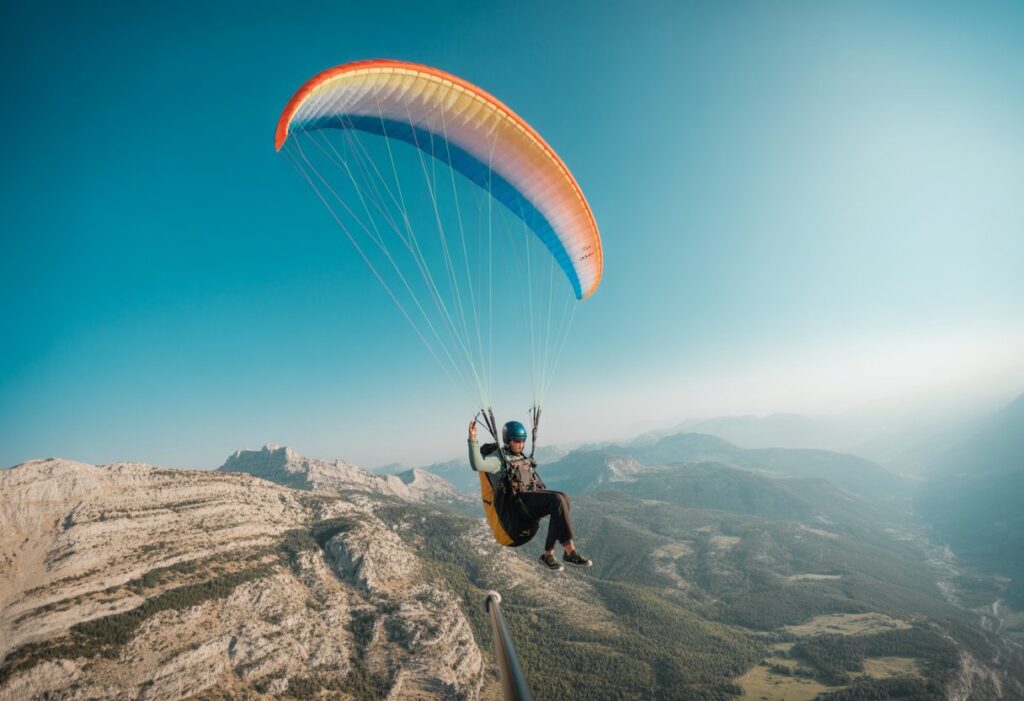 A paraglider pilot in flight leaning into a turn over mountains under a blue sky.
