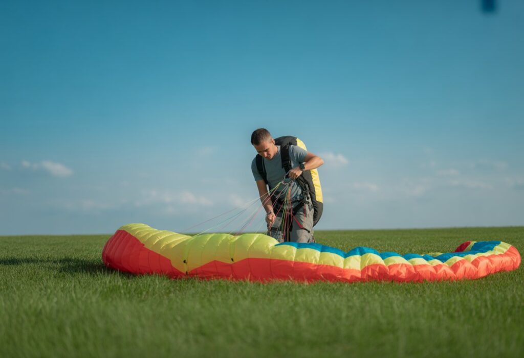 A person preparing to inflate a colorful paraglider wing on a grassy field under a blue sky.