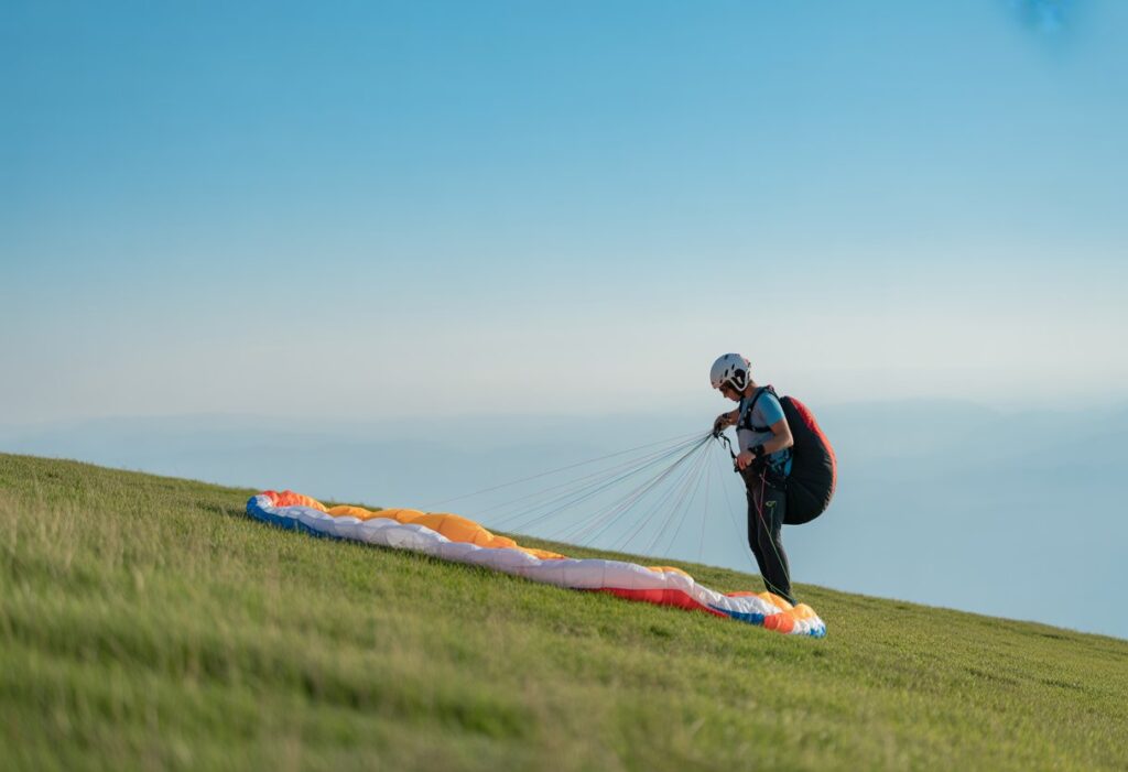 A person preparing a paraglider wing for inflation on a grassy hillside under a clear sky.