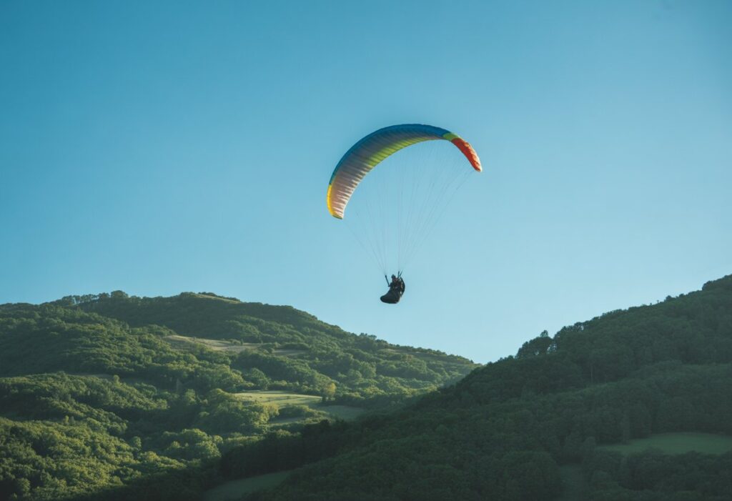 A paraglider circling in the sky above green mountains under a clear blue sky.