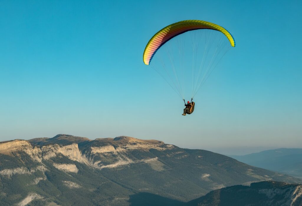A paraglider pilot flying over a mountainous landscape with a bright canopy against a blue sky.