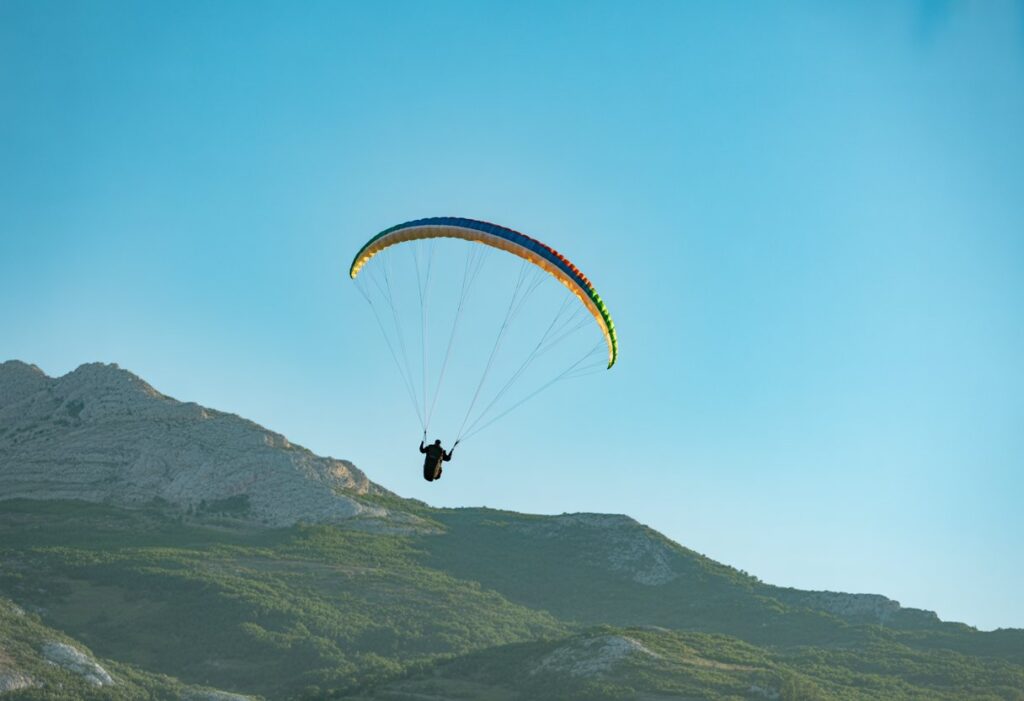 A paraglider performing aerobatic maneuvers high above a mountainous landscape under a clear blue sky.