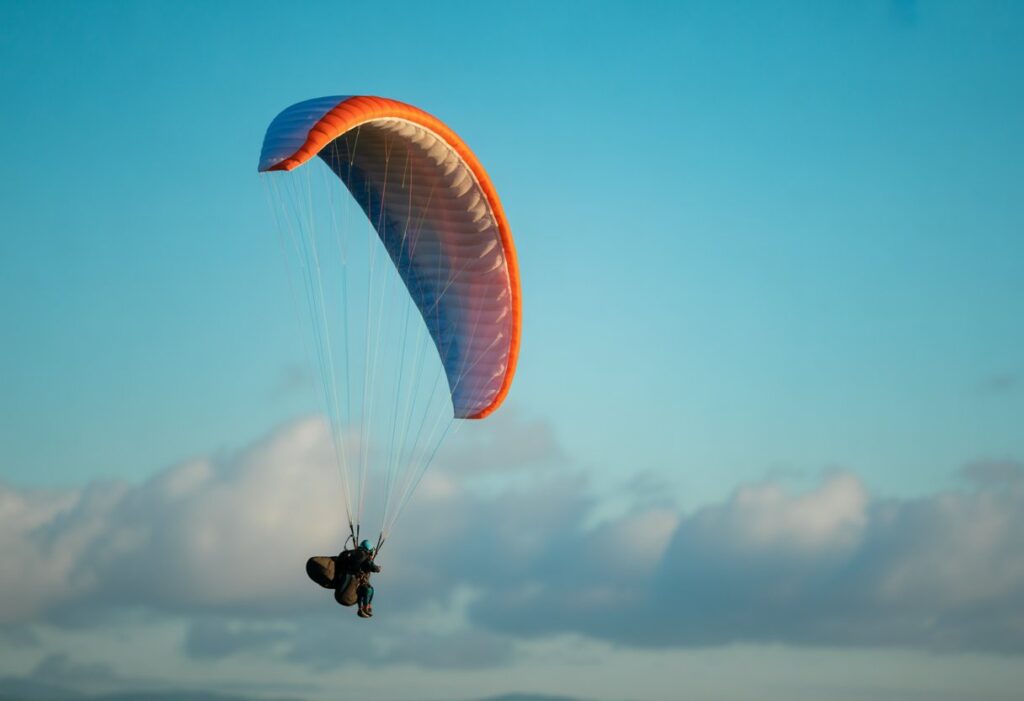 An acro paragliding wing flying in a clear blue sky with clouds in the distance.


