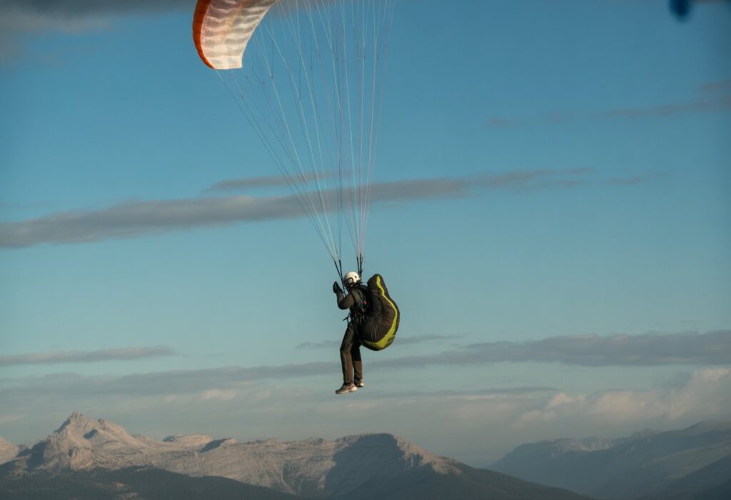 A paraglider in mid-air deploying a reserve parachute with mountains and sky in the background.