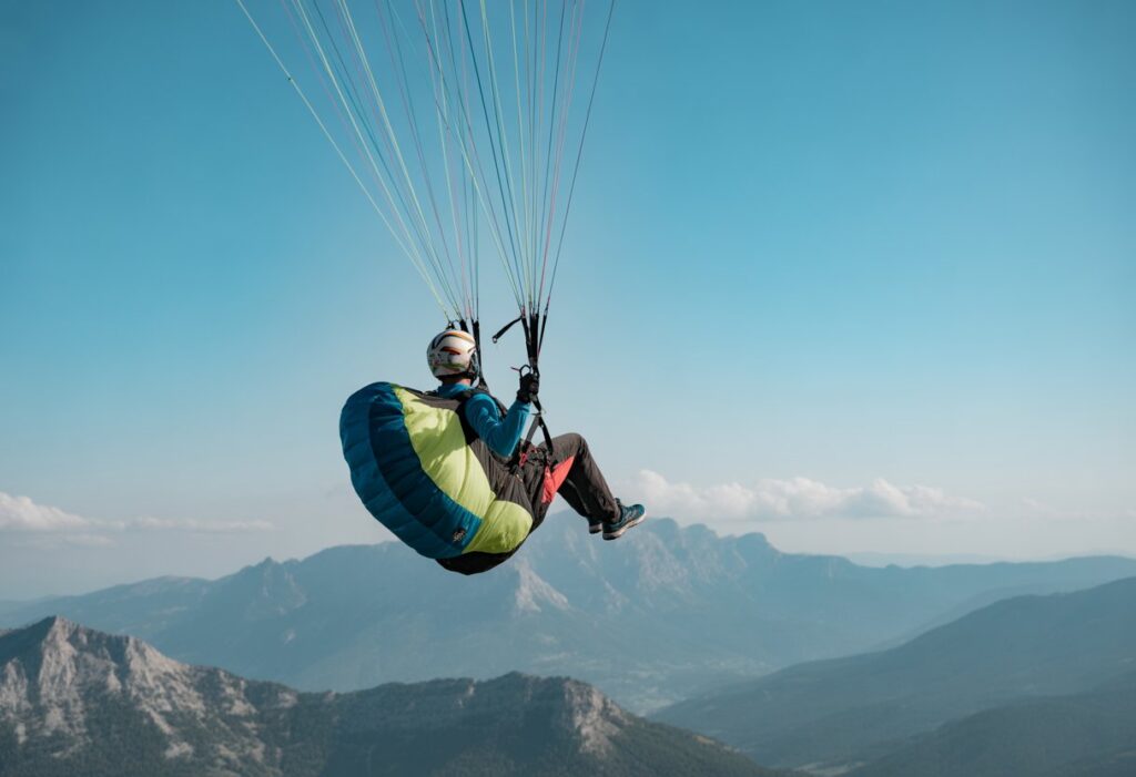 A paraglider in mid-air deploying a reserve parachute with mountains and blue sky in the background.