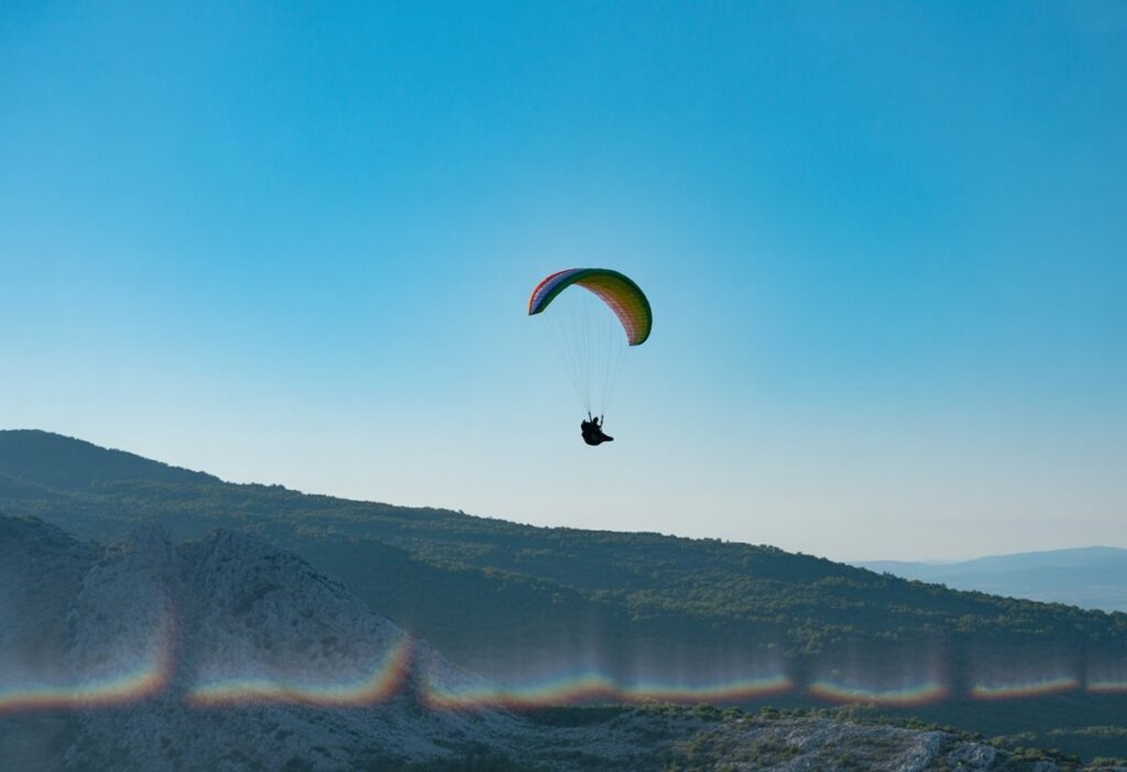 A paraglider flying above mountains on a sunny day with clear blue sky and visible heat waves rising from the terrain.