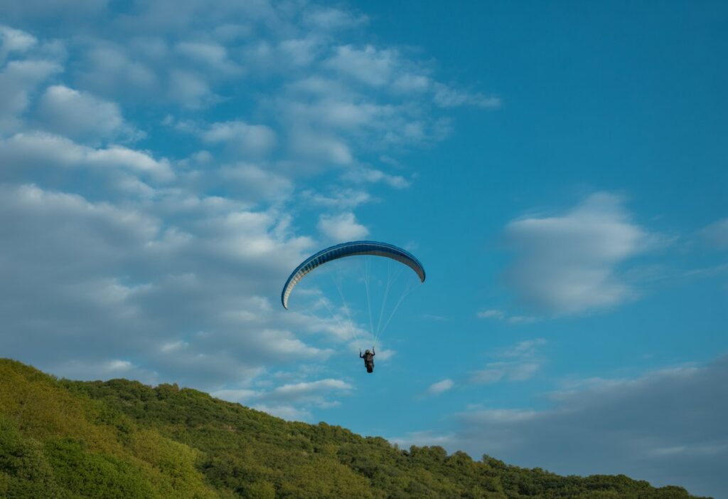 A paraglider flying over green hills beneath a blue sky with scattered white clouds showing thermal updrafts.