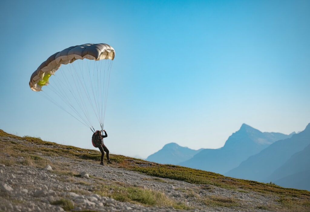 A person landing a parachute on a rocky mountain slope under a clear sky, with distant alpine peaks in the background.