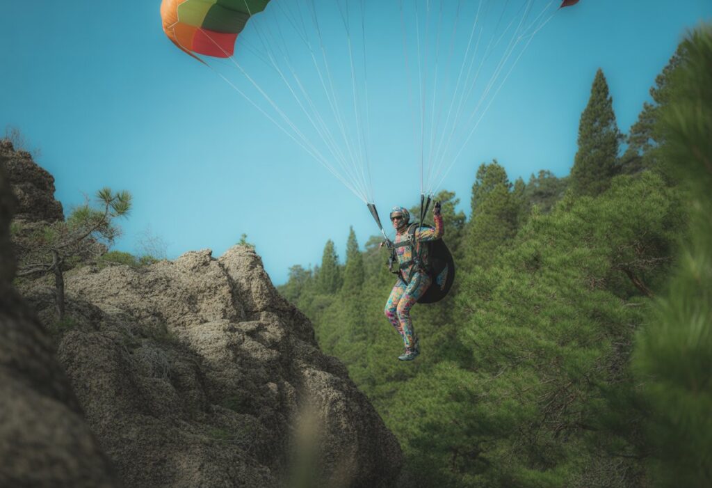 A parachutist in a colorful suit landing near rocky mountainous terrain, surrounded by pine trees under a clear blue sky.